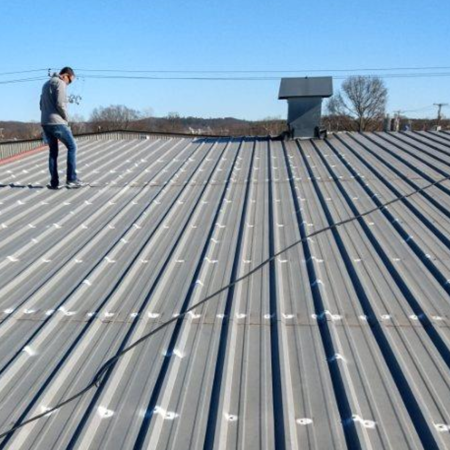Worker on a metal roof inspecting seams with a tool, with ventilation units and a clear sky in the background