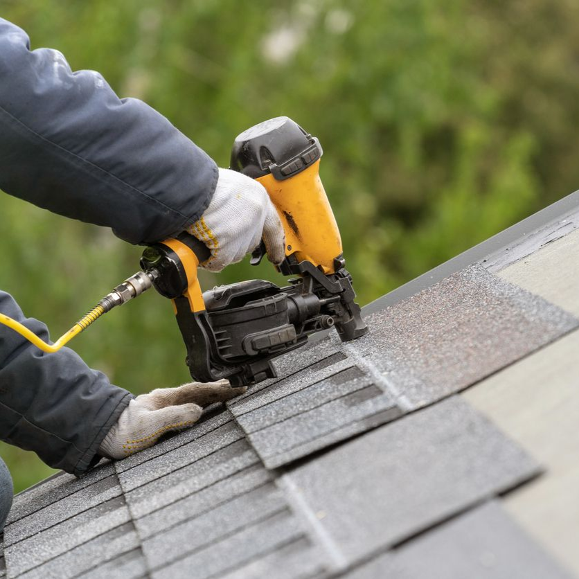 Worker using a yellow nail gun to install gray shingles on a roof