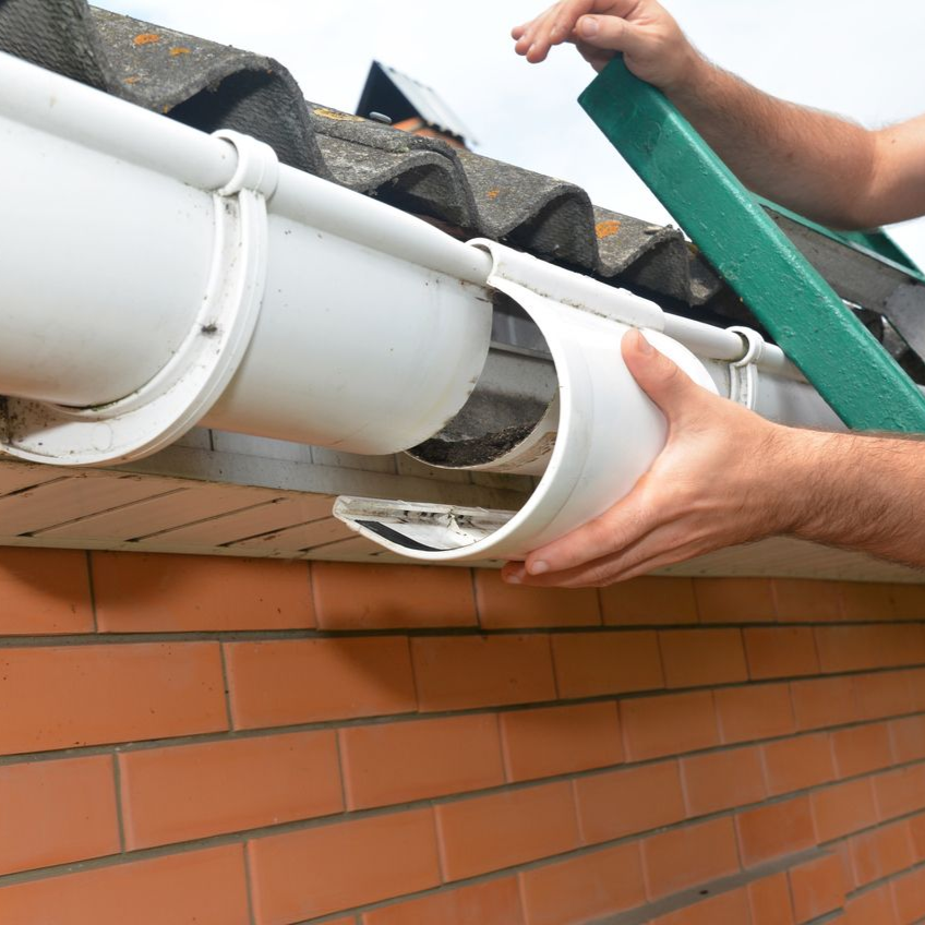 Hands installing a white gutter on a brick house roofline