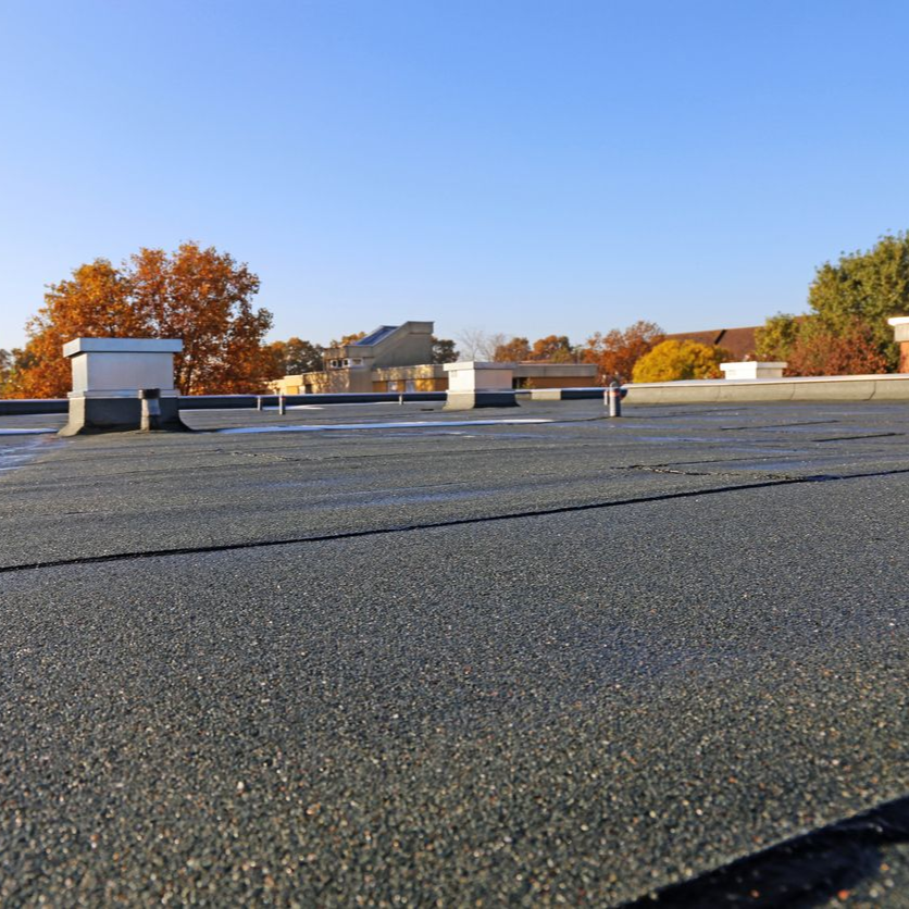 Flat rooftop with gravel surface and white vents under a clear blue sky, autumn trees in the background