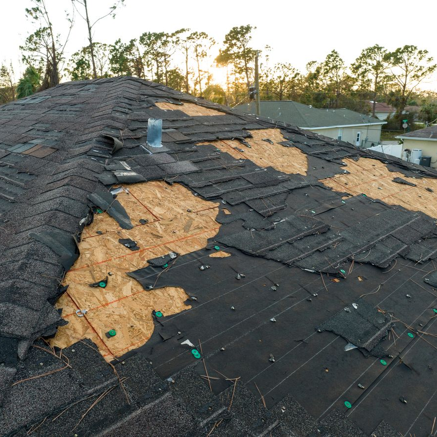 Damaged shingle roof with several torn sections exposing underlayment at sunset