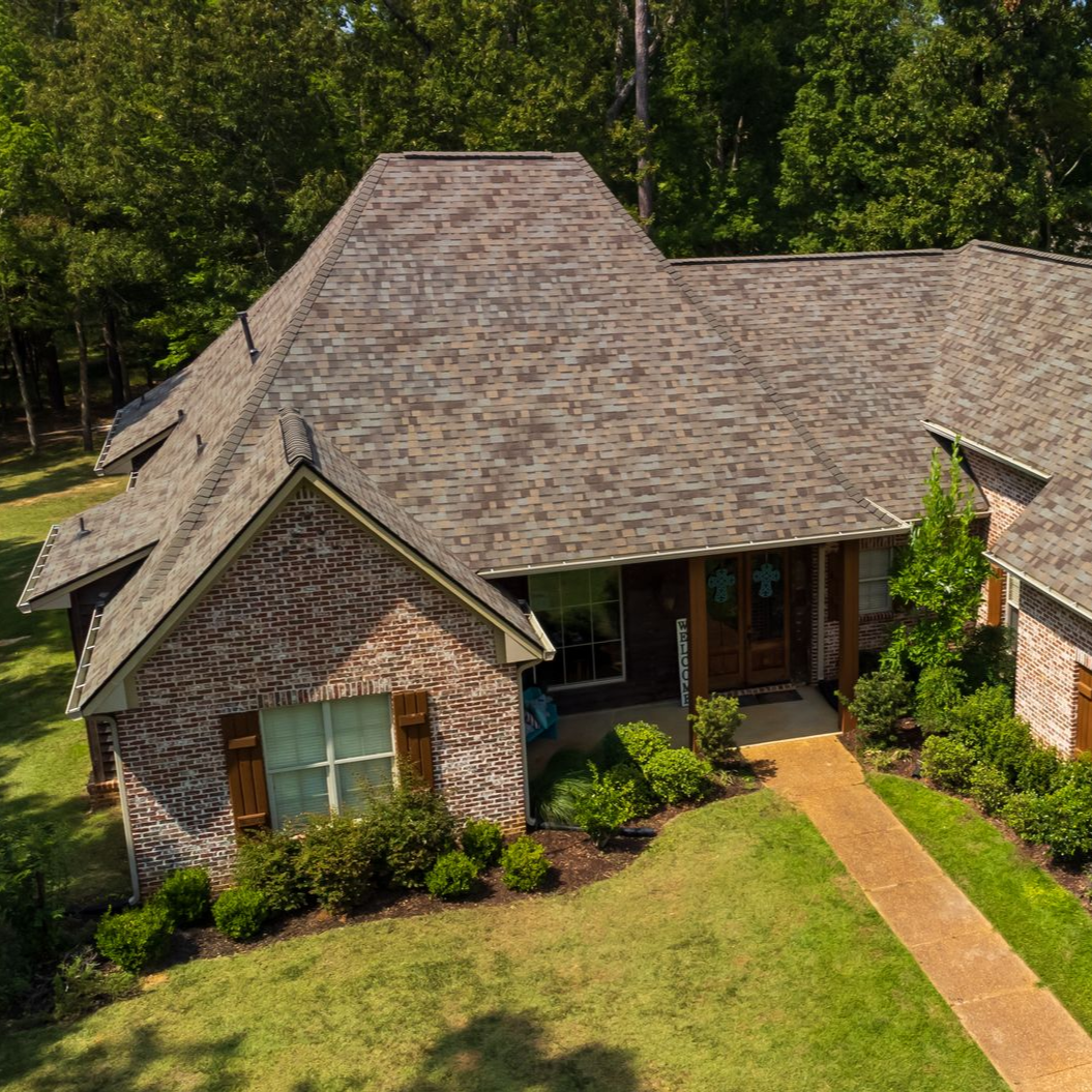 Suburban brick house with a dark shingled roof, front lawn, and walkway surrounded by trees