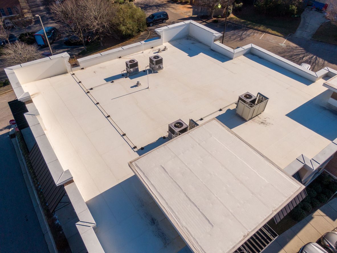 Aerial view of a flat white rooftop with vents and HVAC units, surrounded by trees and buildings.