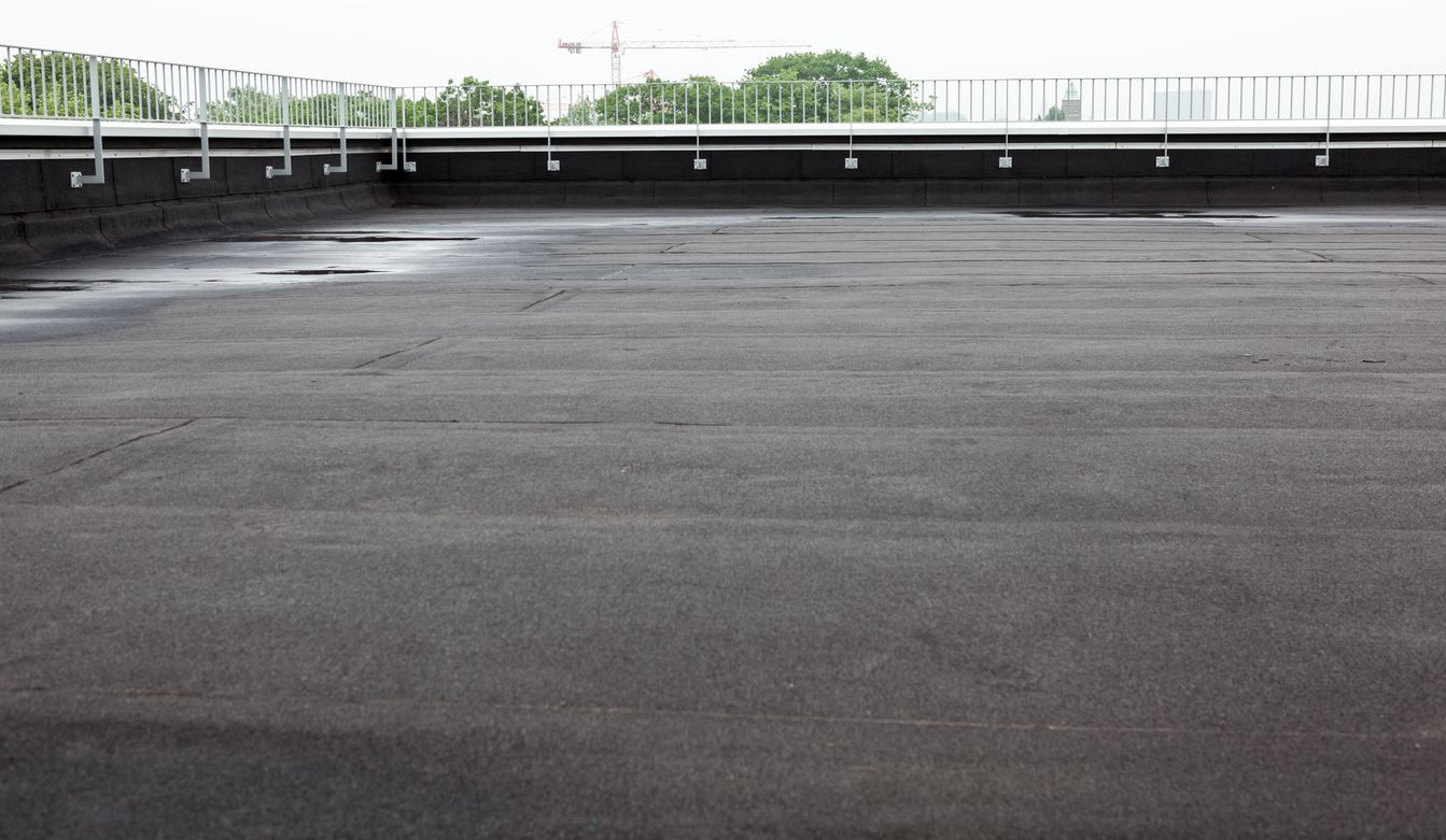 Empty paved rooftop parking lot with a dark surface and a low railing against a green treeline.