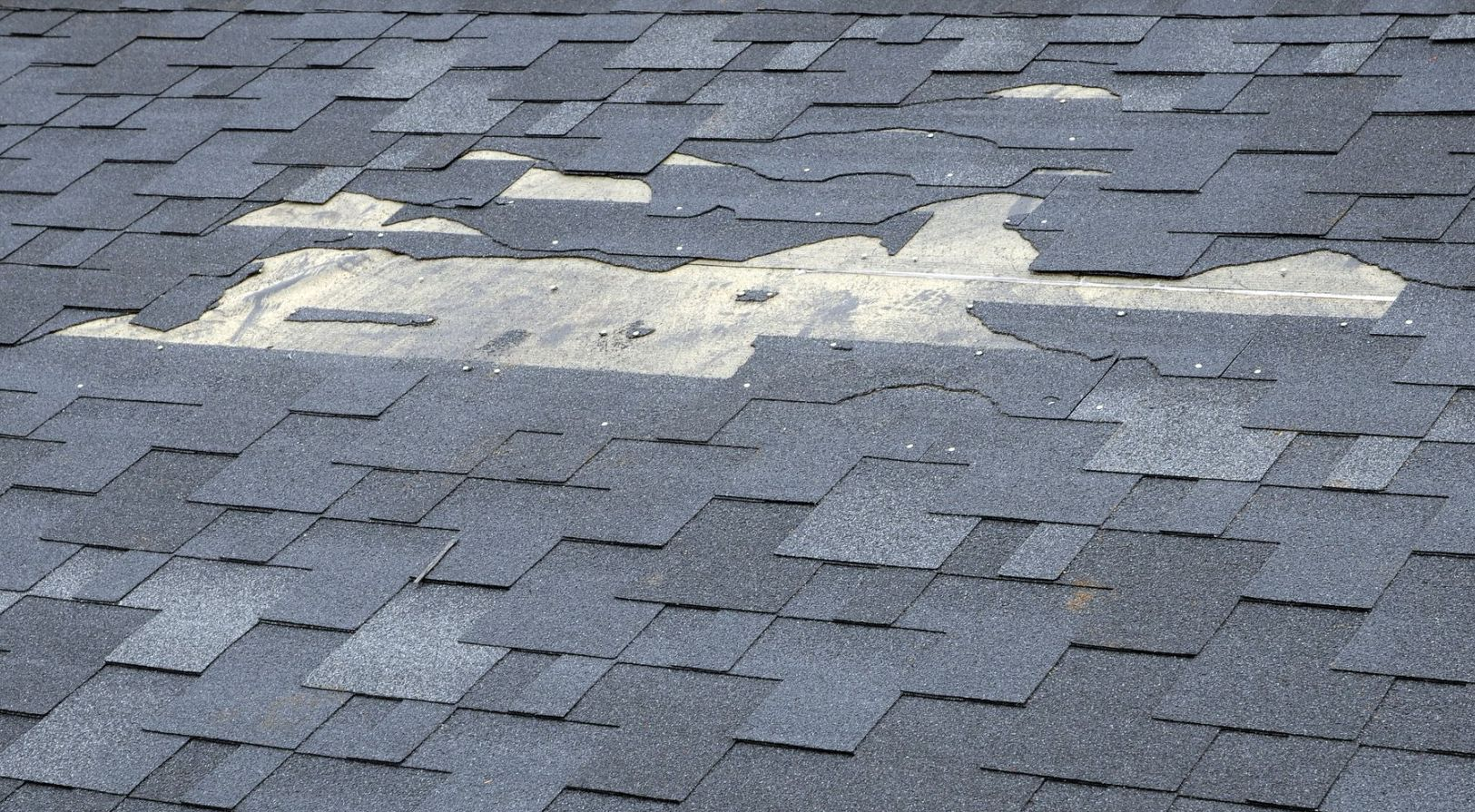 Damaged gray roof shingles with a large patch of missing material exposing lighter underlayment