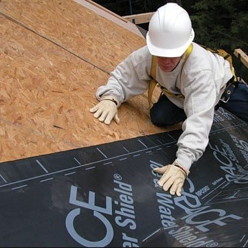 Worker installing black roof underlayment on a sloped roof, wearing a white hard hat and gloves
