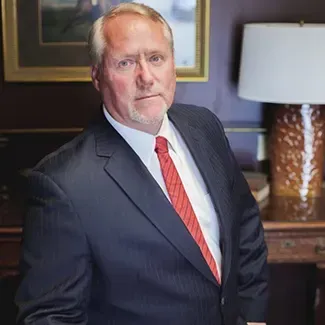 Man in a dark suit and red tie standing in an office beside a lamp and framed artwork