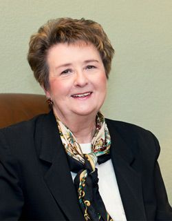 Smiling person in a black blazer and patterned scarf, seated against a plain beige background
