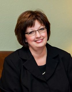 Smiling woman in glasses and a black blazer seated against a beige wall.