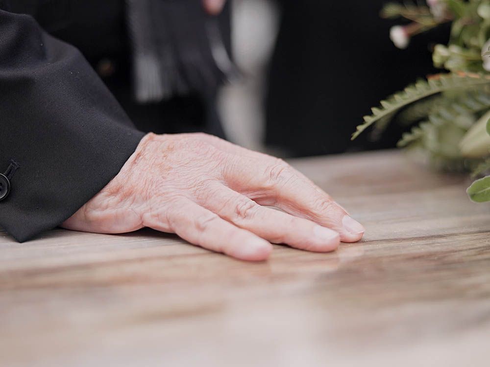 A hand resting on a wooden table beside a small plant, with a dark sleeve in the background