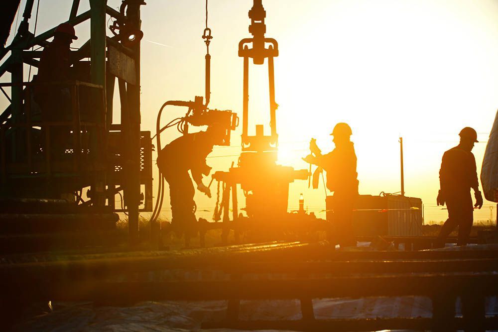 Silhouetted workers on an industrial site at sunset with drilling equipment and bright orange light