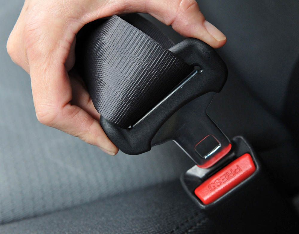 Hand pulling a black seat belt buckle near a red release button in a car seat.