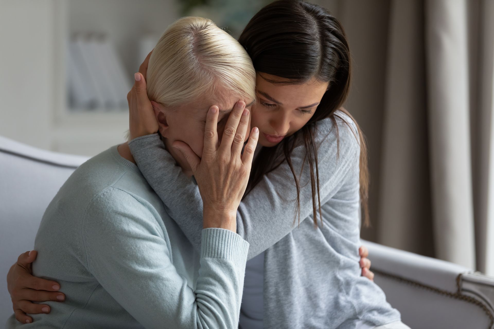 Two people sitting closely, one comforting the other who is covering their face, in a quiet room