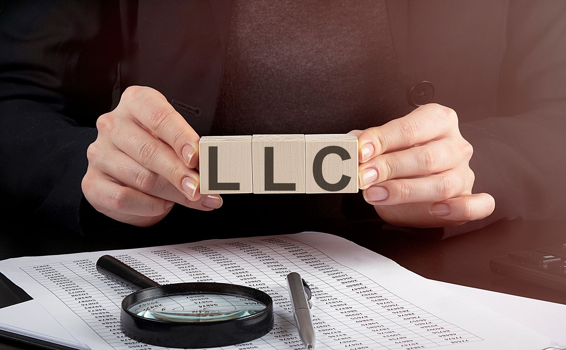 Hands holding letter blocks spelling “LLC” above paperwork with a magnifying glass and pen
