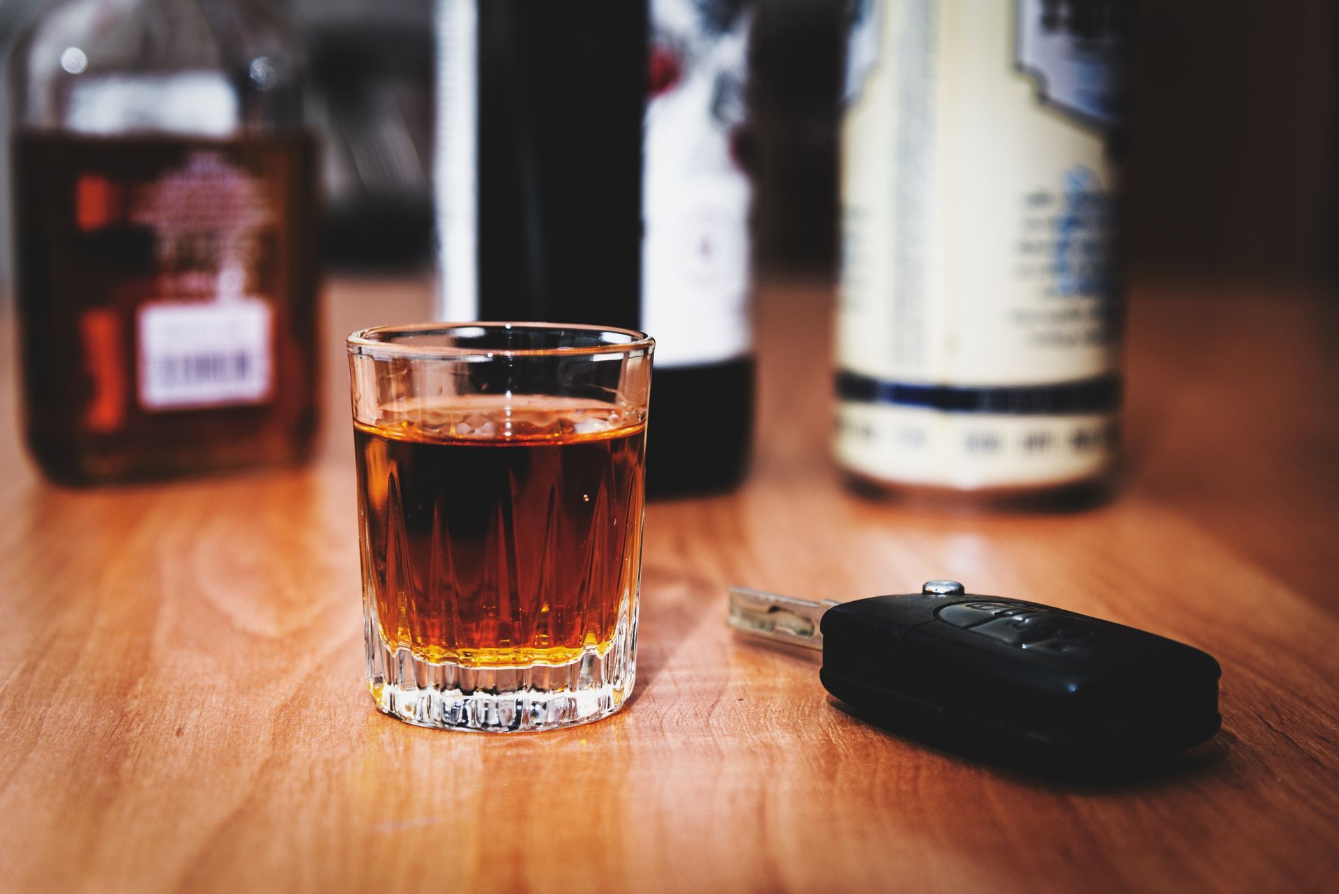 A shot glass of amber liquor on a wooden table with blurred bottles in the background and keys beside it
