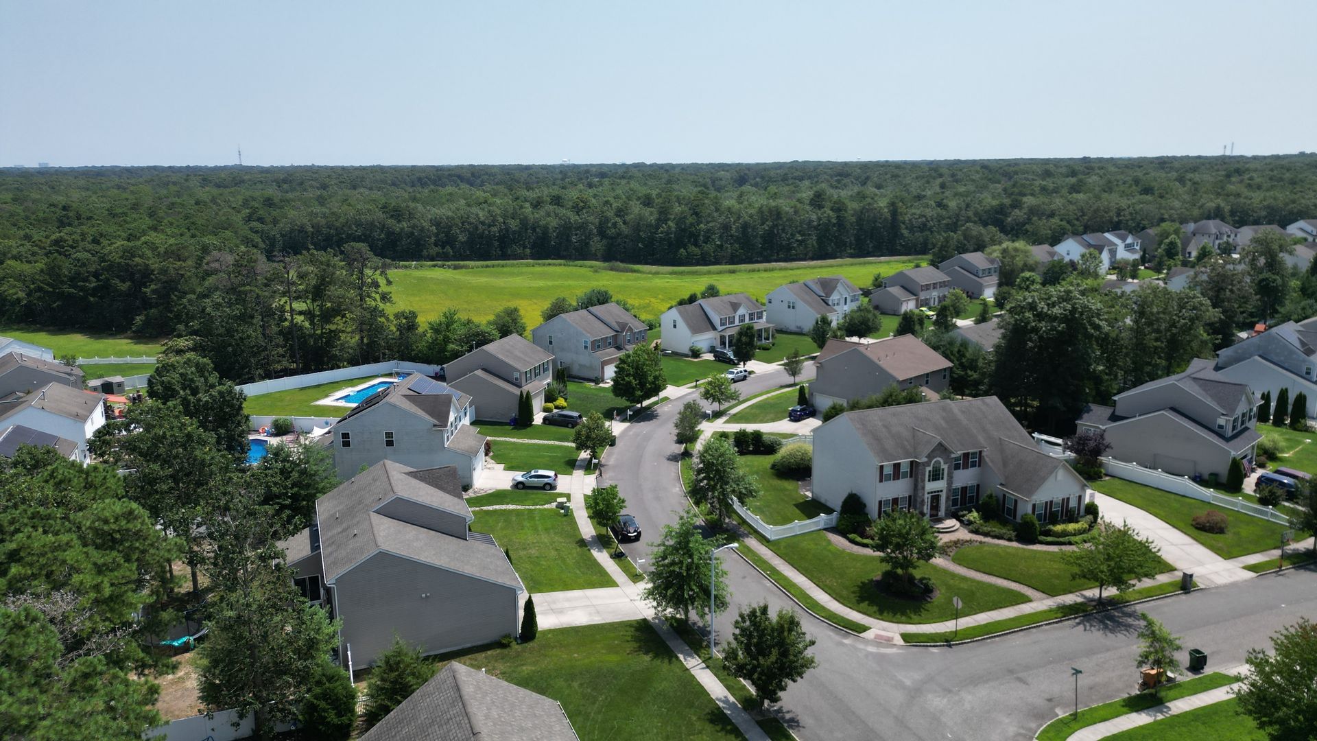 Aerial view of a suburban neighborhood with gray-roofed houses, winding streets, and surrounding trees.