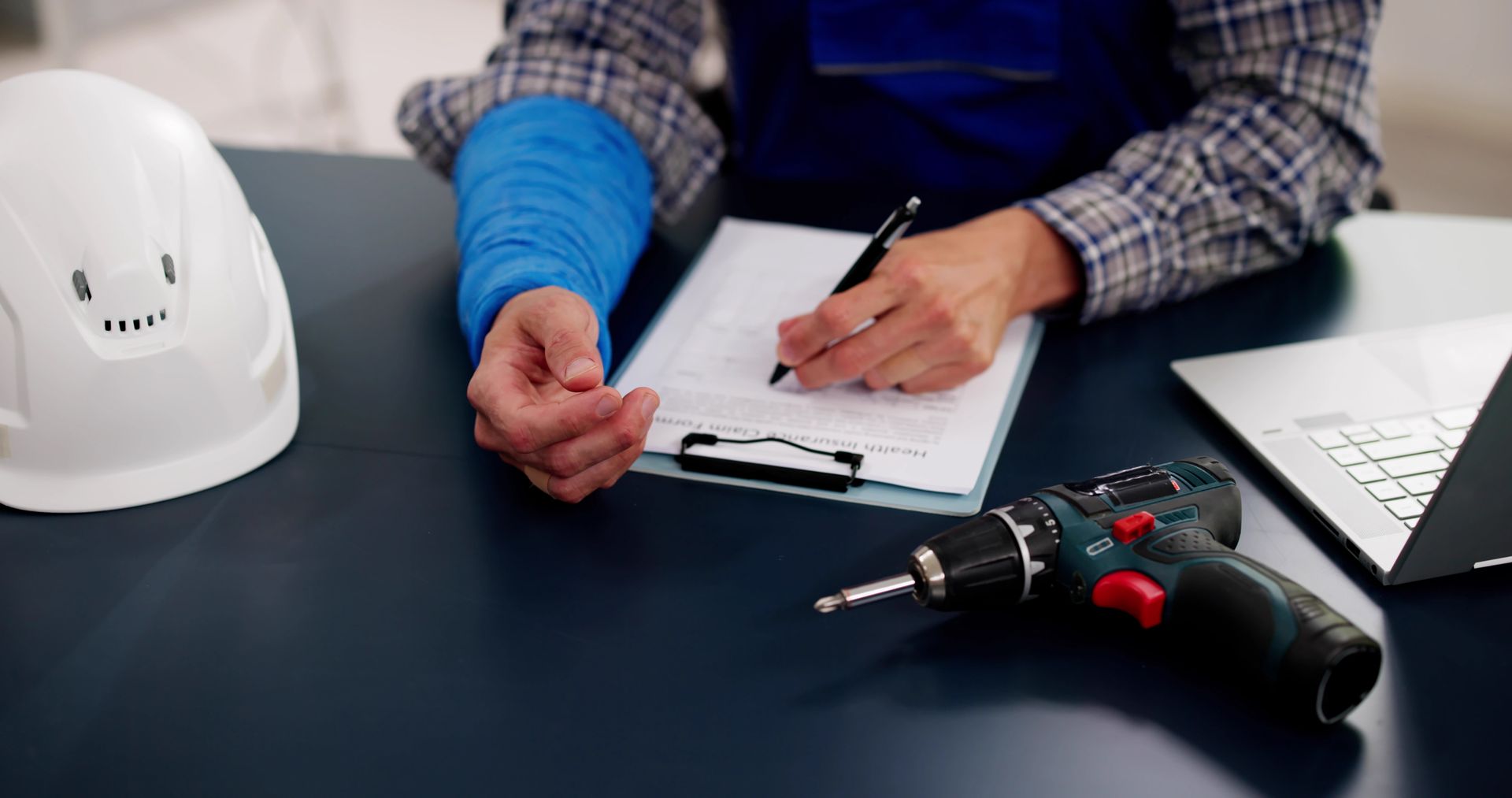 Person writing on a clipboard beside a drill, laptop, and white hard hat on a desk.
