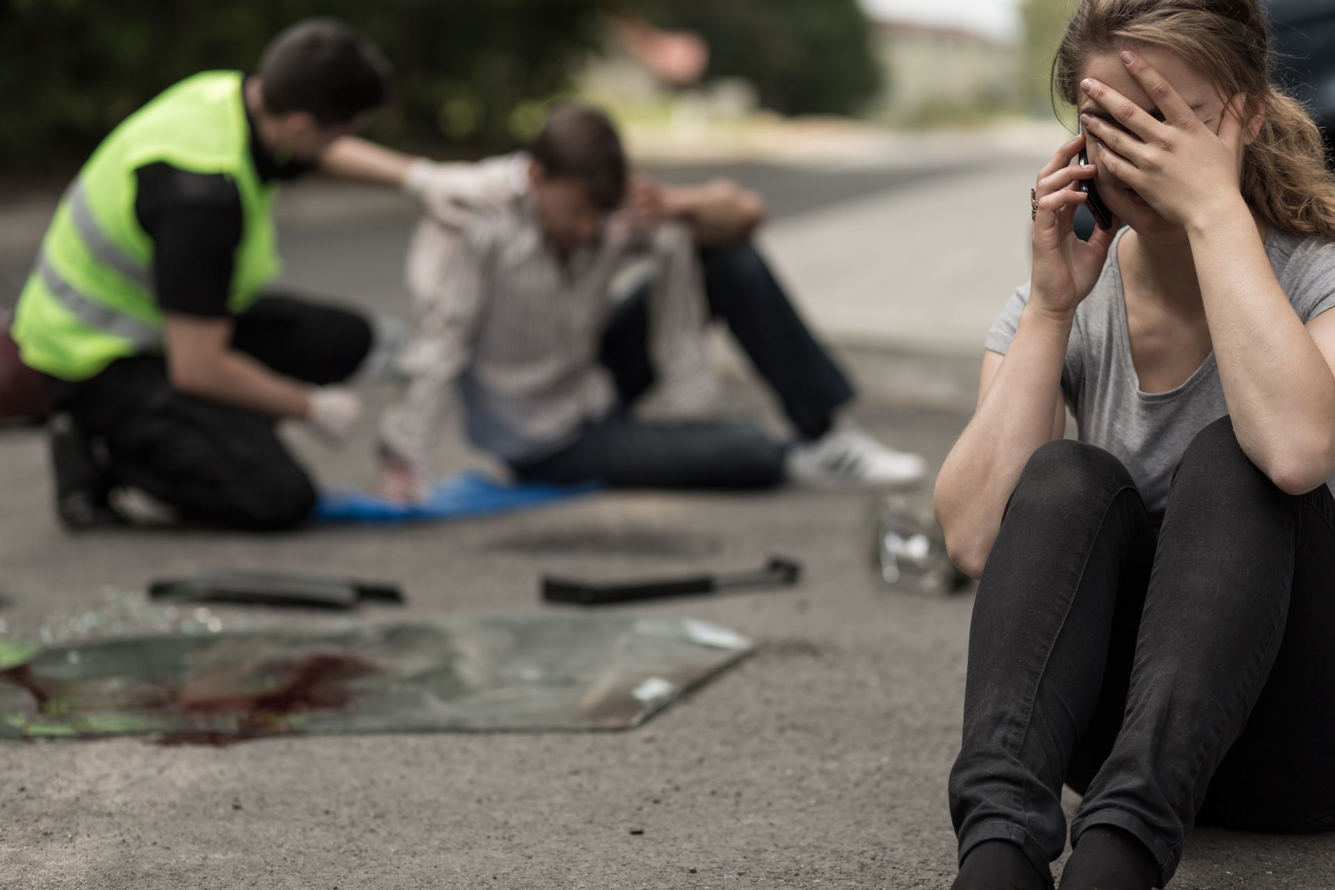 Woman sitting on road, covering her face, while responders attend to an injured person in the background