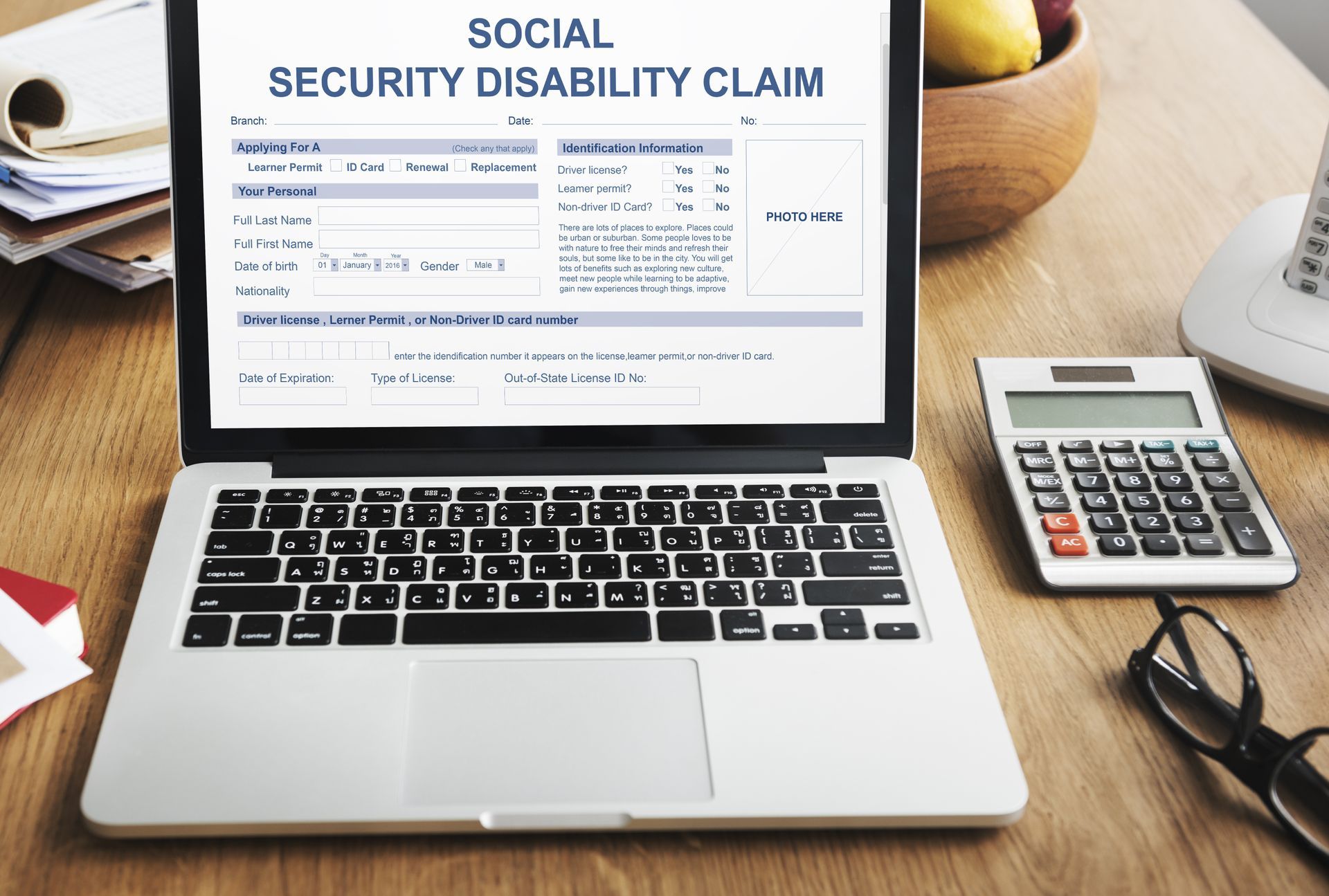 Laptop displaying a Social Security Disability Claim form on a desk with calculator, glasses, and paperwork