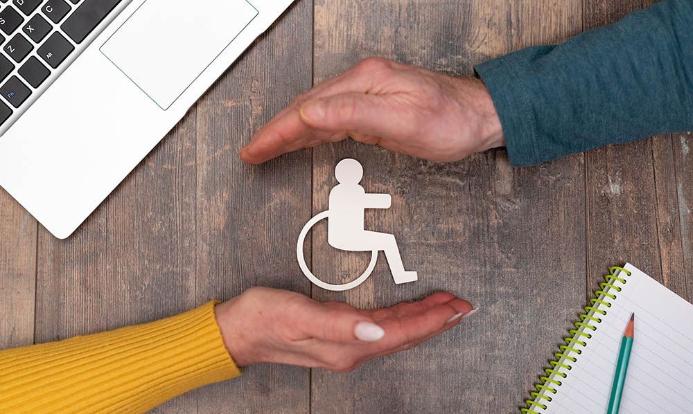 Two hands cupping a wheelchair icon on a wooden desk with a laptop and notebook nearby