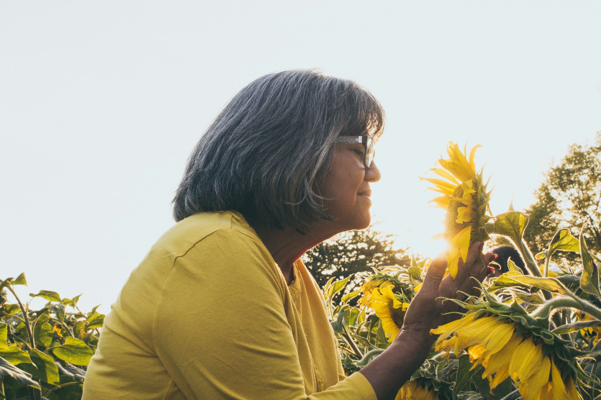 A woman in a yellow shirt is smelling a sunflower in a field.