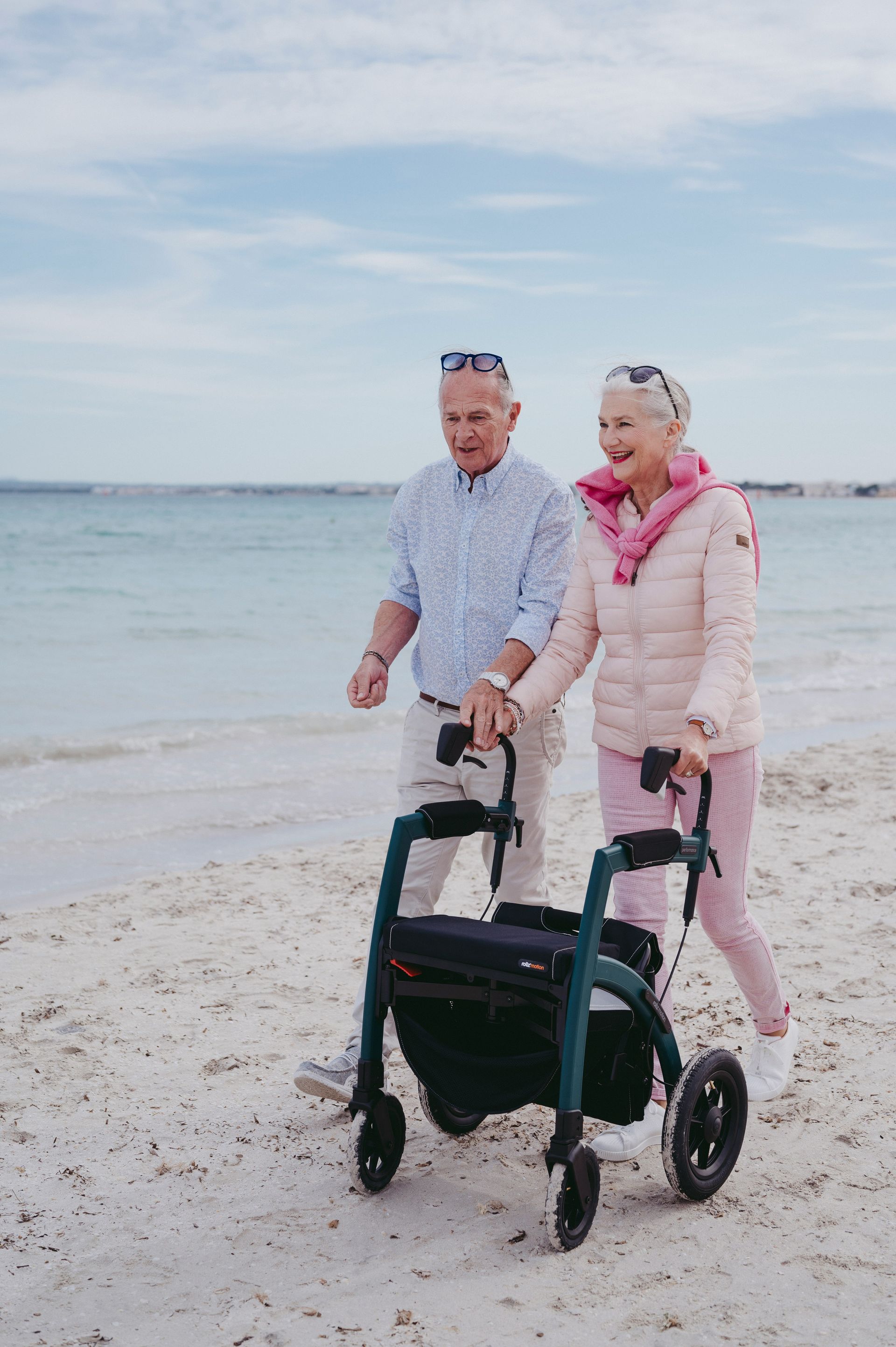 An elderly couple is walking on the beach with a walker.