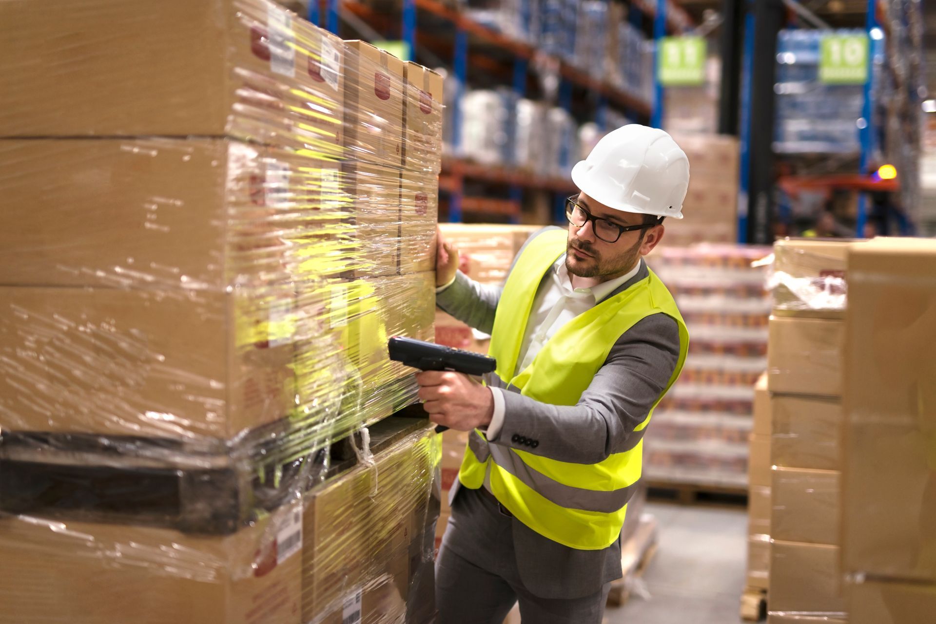 Worker in a hard hat and safety vest scanning palletized boxes in a warehouse.