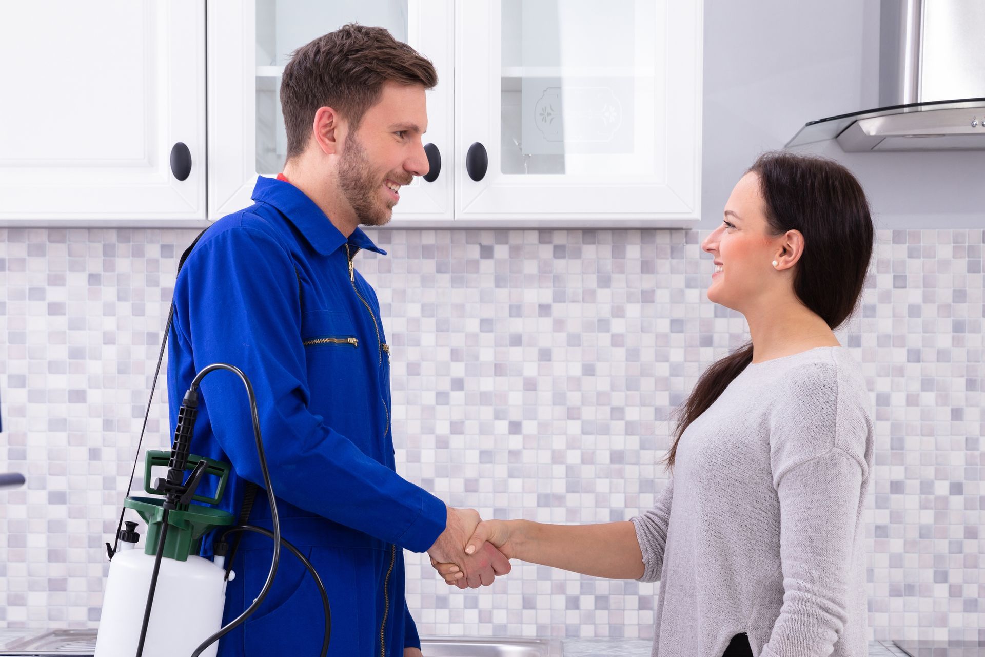 A pest control worker, wearing a blue uniform, shaking hands with a woman, in her kitchen.