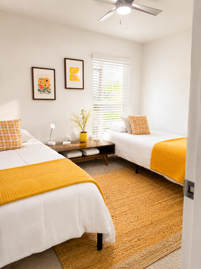 Bright twin bedroom with white bedding, mustard blankets, woven rug, and a small nightstand between beds