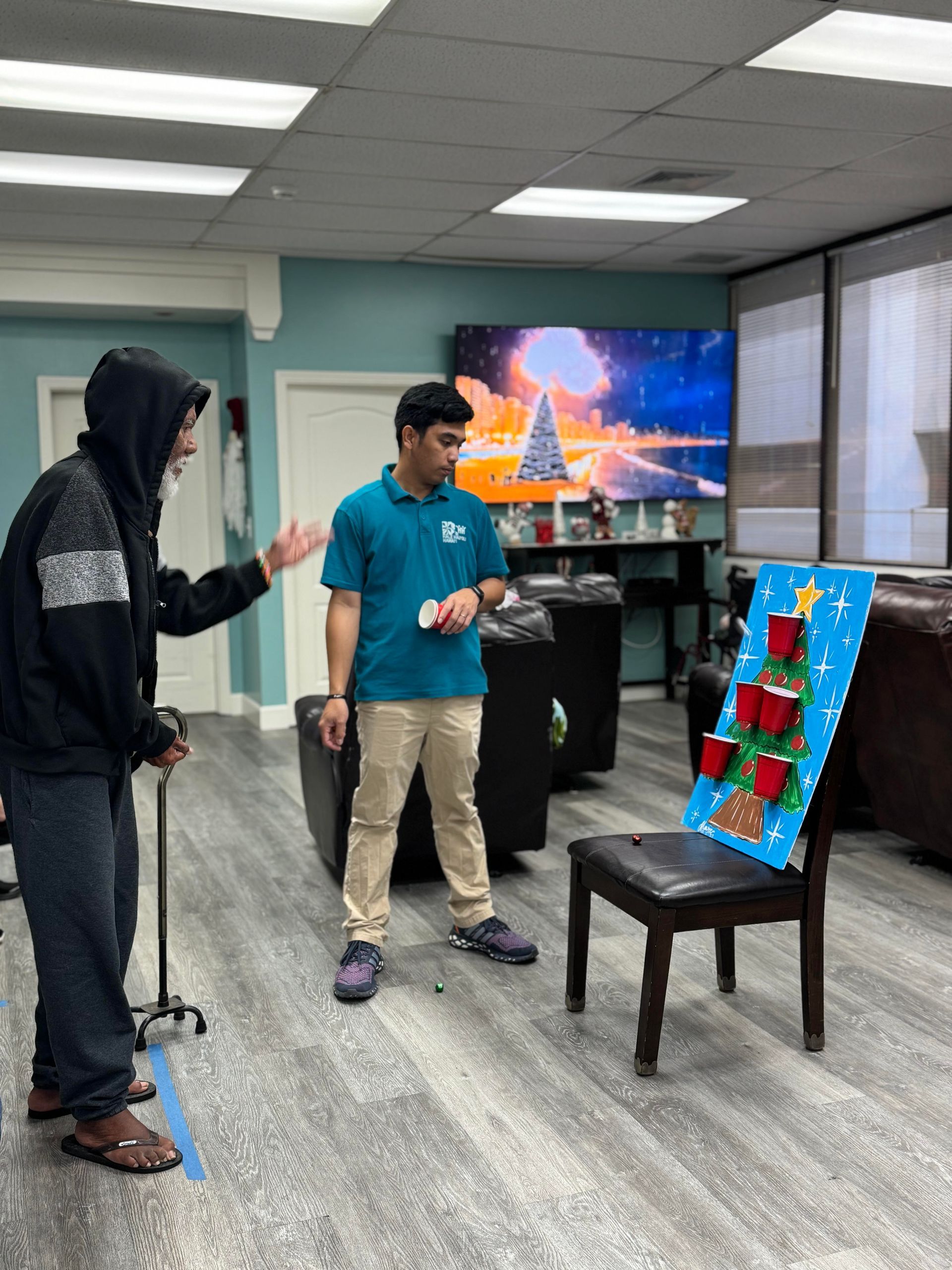 A male caregiver assists a senior man playing a holiday cup game in an activity room.