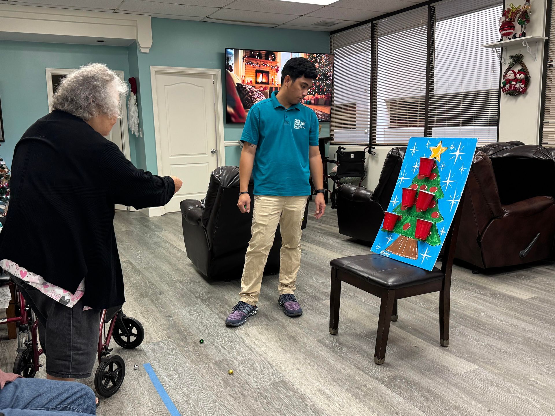 A senior woman tosses a ball into a Christmas tree cup game while a male staff member watches.