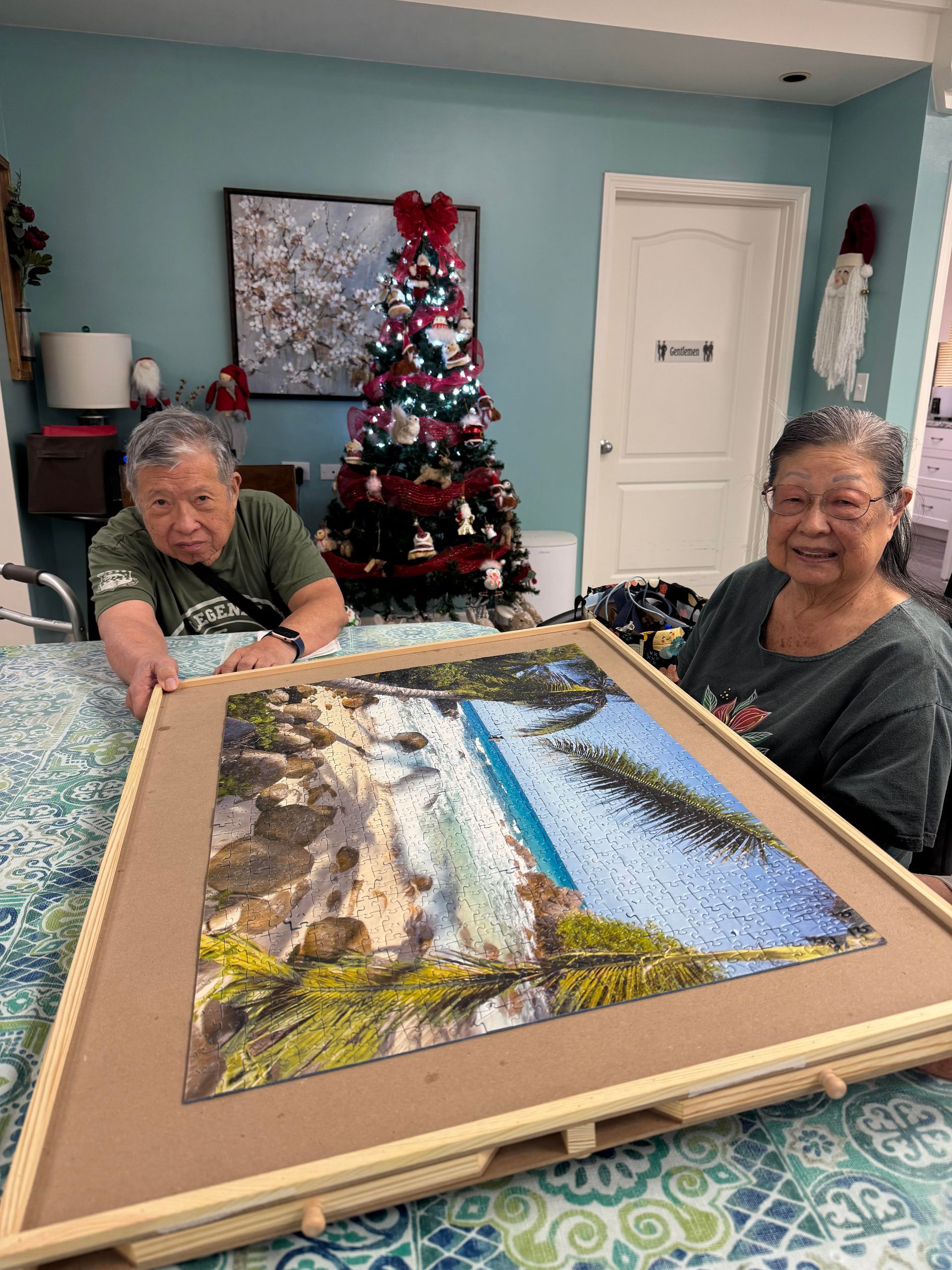 An elderly man and woman smile while showcasing a large, completed tropical beach jigsaw puzzle.