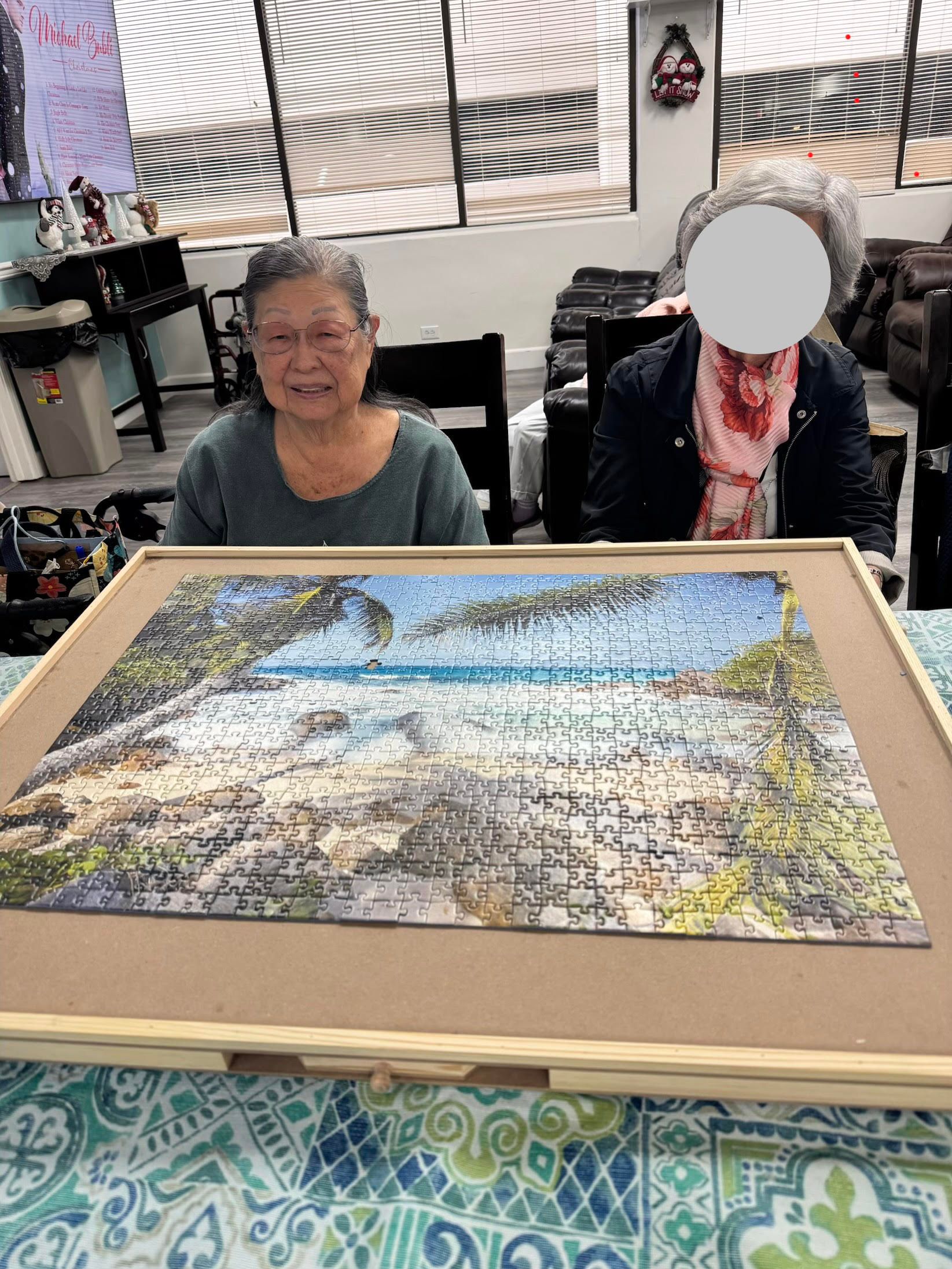 Two senior women sit behind a large completed jigsaw puzzle of a tropical beach landscape.