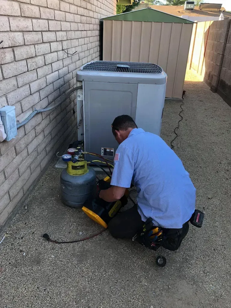 Technician servicing an outdoor HVAC unit beside a brick wall and fence, with tools on the ground.
