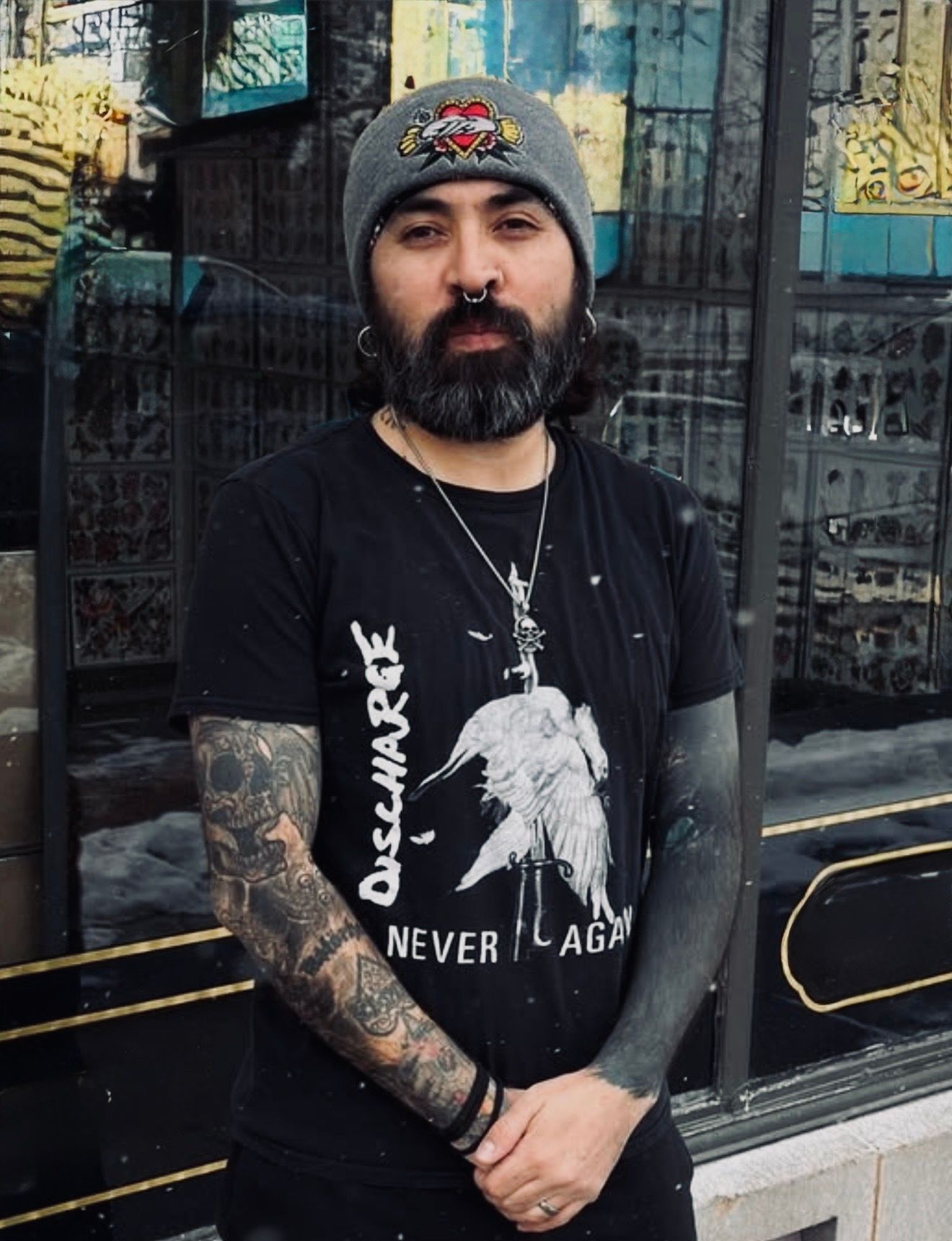 Man with beard and tattoos wearing a gray beanie and black graphic T-shirt, standing in front of a storefront.