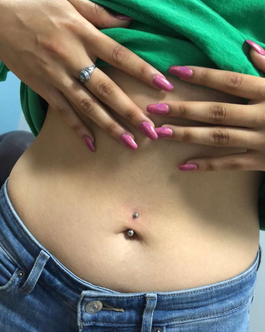 Person's stomach with belly button piercing, hands on waist, wearing jeans and green top.