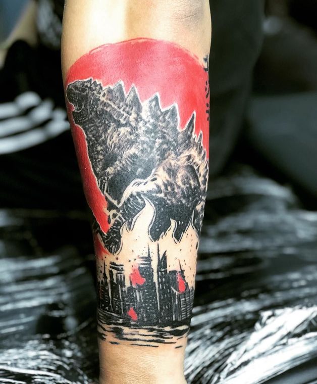 Godzilla tattoo on forearm, black silhouette with a red sun, cityscape below.