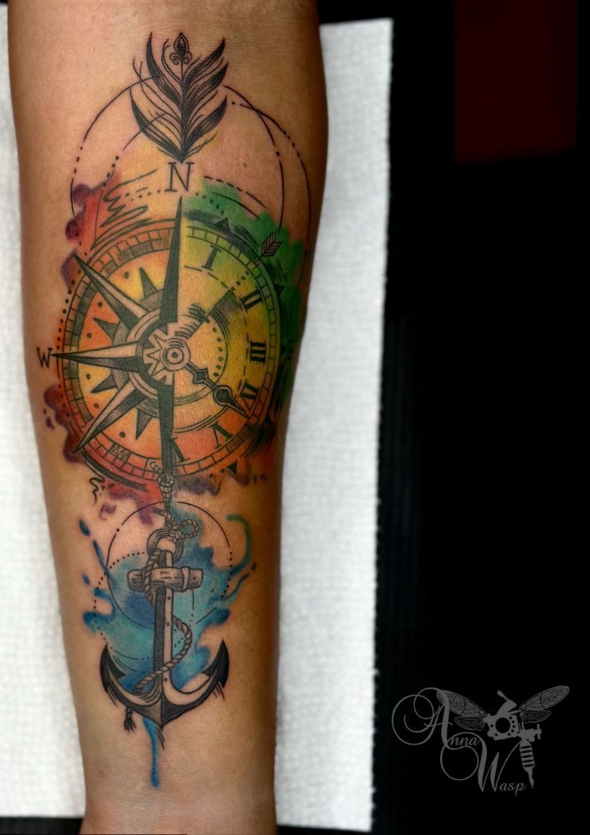 Tattoo of a compass, anchor, and feather with watercolor effects on an arm; colorful design.
