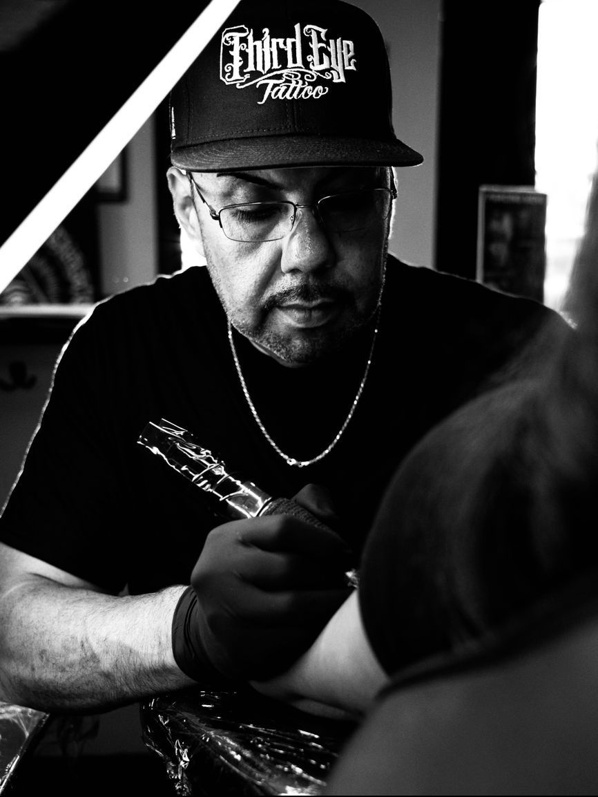 Tattoo artist wearing a hat and glasses, working on a client's arm. Black and white.