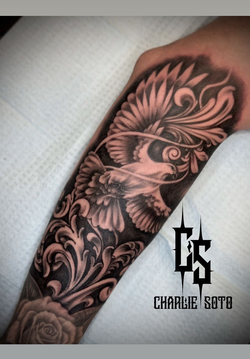 Tattoo of a dove with spread wings amid ornate, swirling floral designs, on an arm. Black and gray shading.