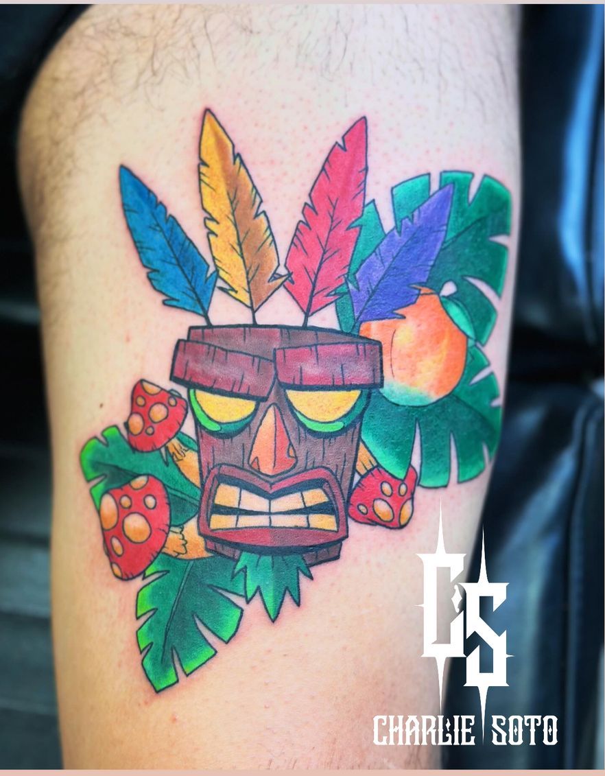 Tattoo of a wooden tiki mask with colorful feathers, surrounded by plants and mushrooms, on a leg.