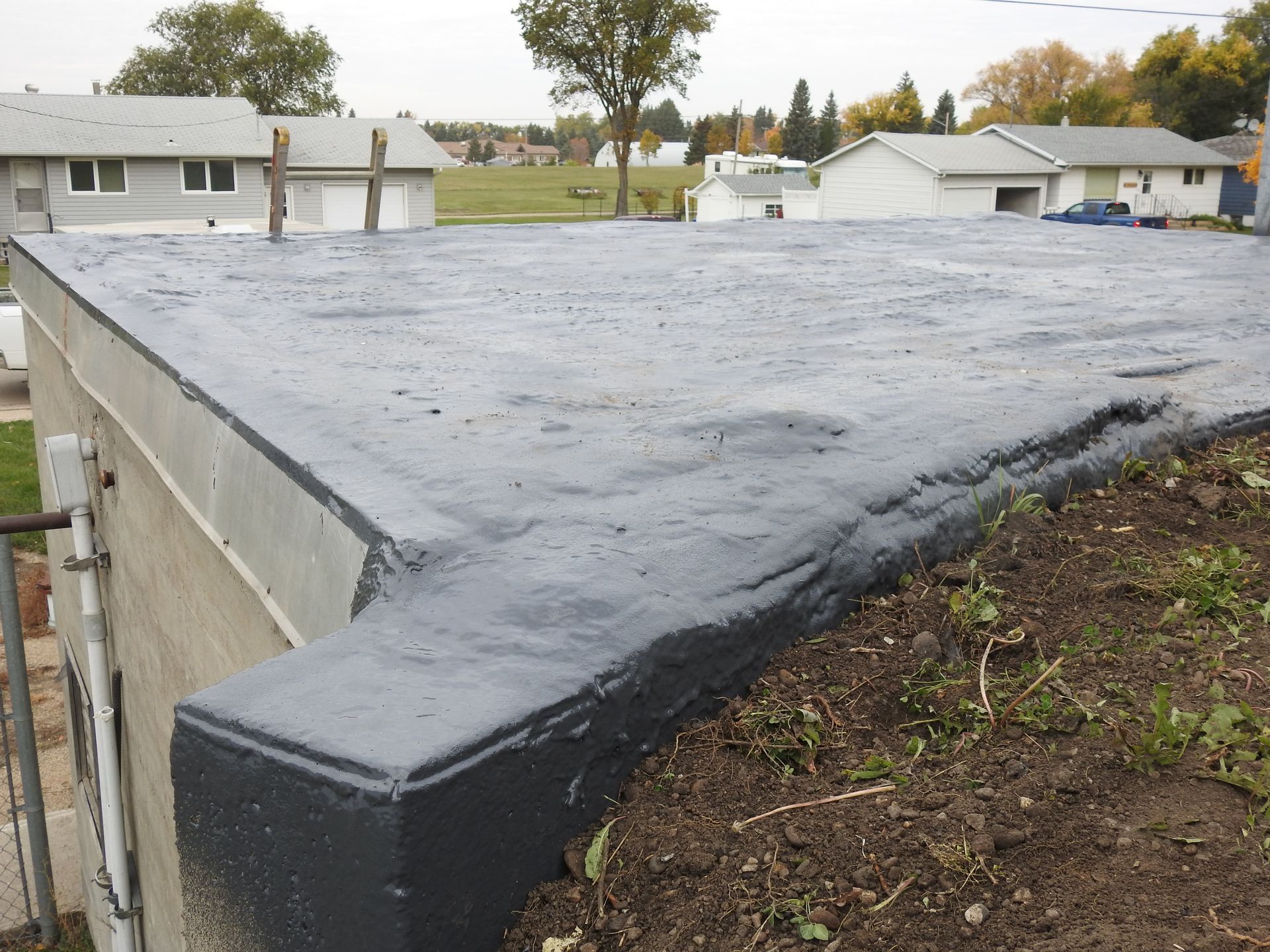 Flat, freshly painted dark grey roof. Concrete edge with dirt and grass in foreground. Houses in background.