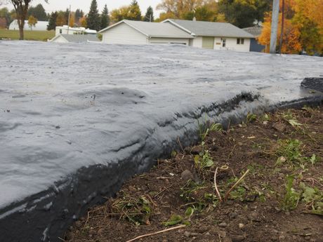Black asphalt surface, possibly a roof, meeting brown dirt with weeds. Houses visible in the background.