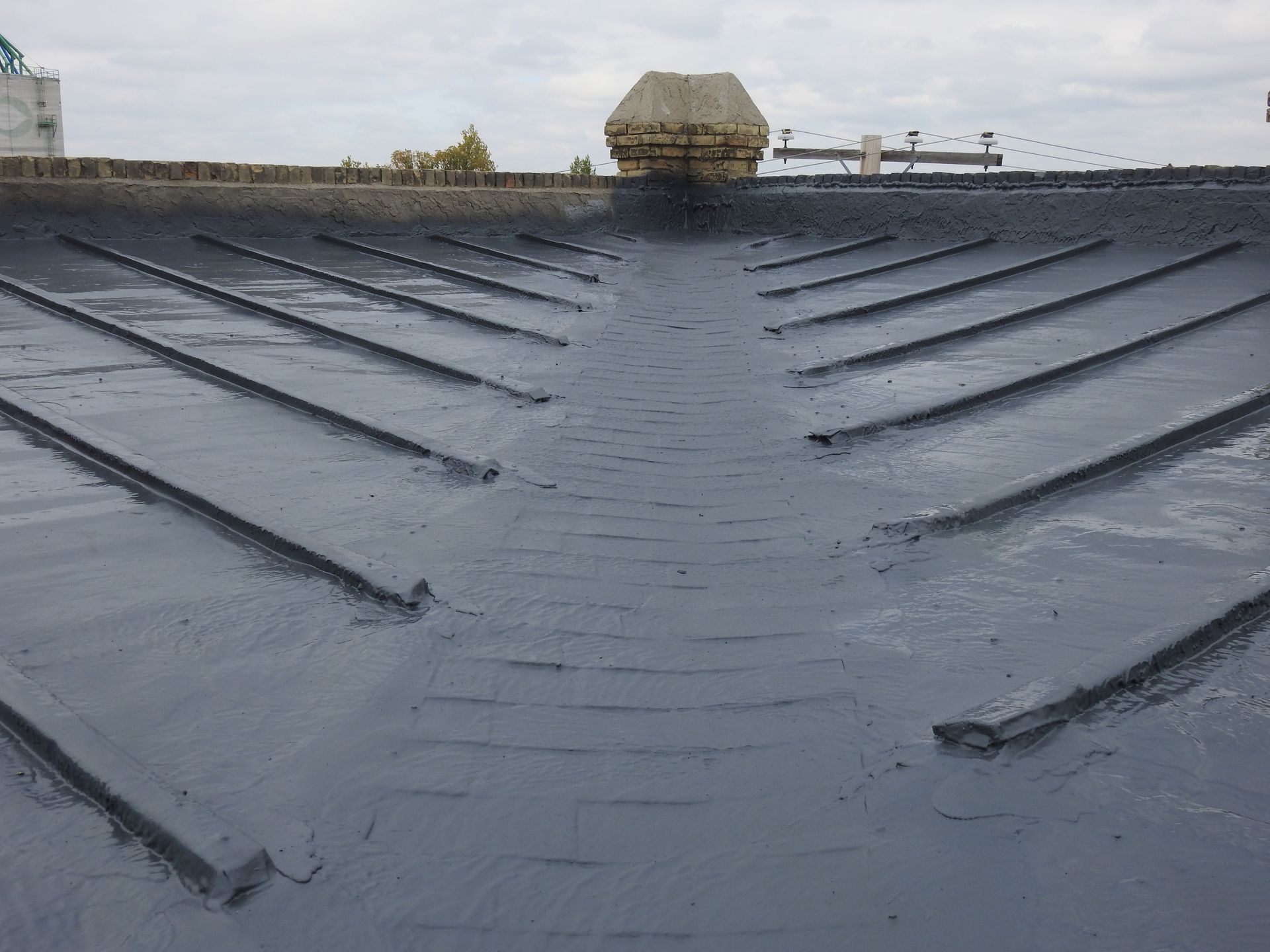 Dark gray roof with angled ridges, a central drainage channel, and a chimney against a cloudy sky.