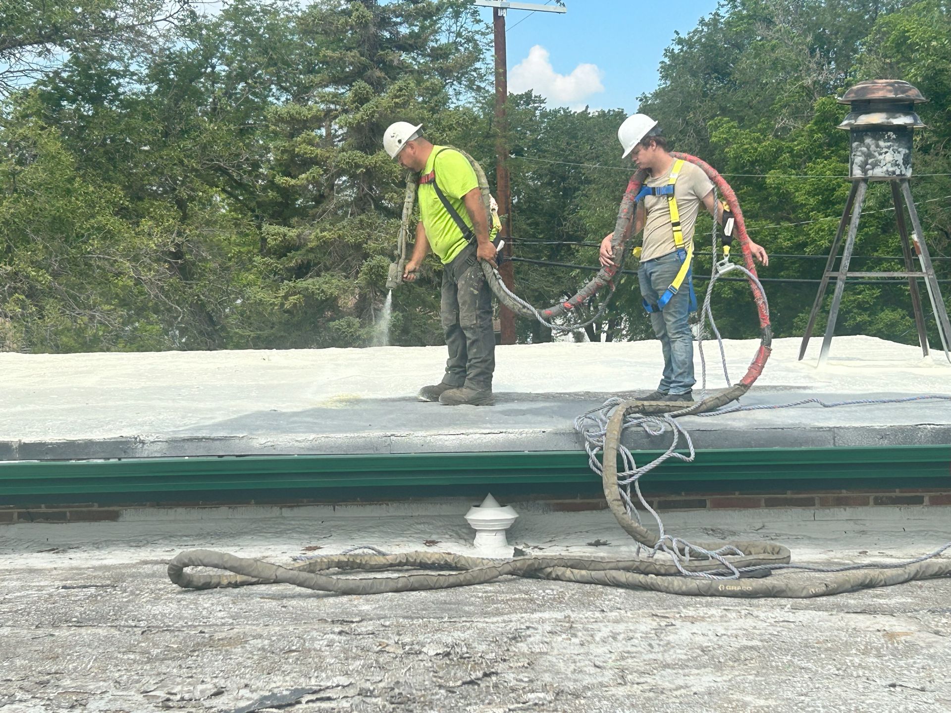 Two workers in safety gear on a rooftop, holding hoses. Trees and a chimney are visible in the background.