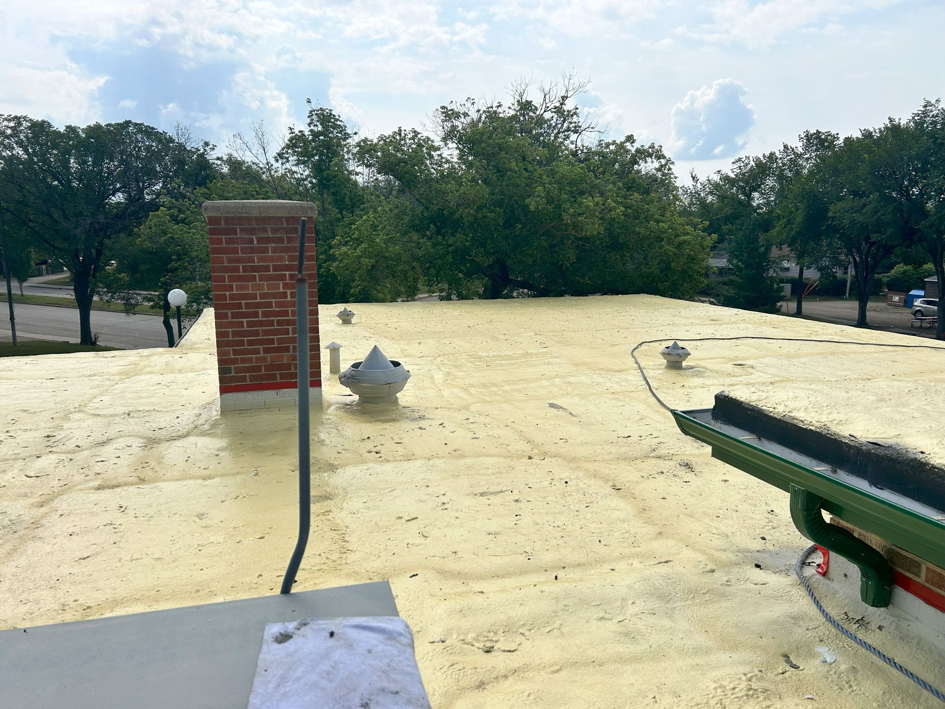 A beige foam-coated rooftop with a brick chimney, three vents, and a gutter, trees in the background under a cloudy sky.