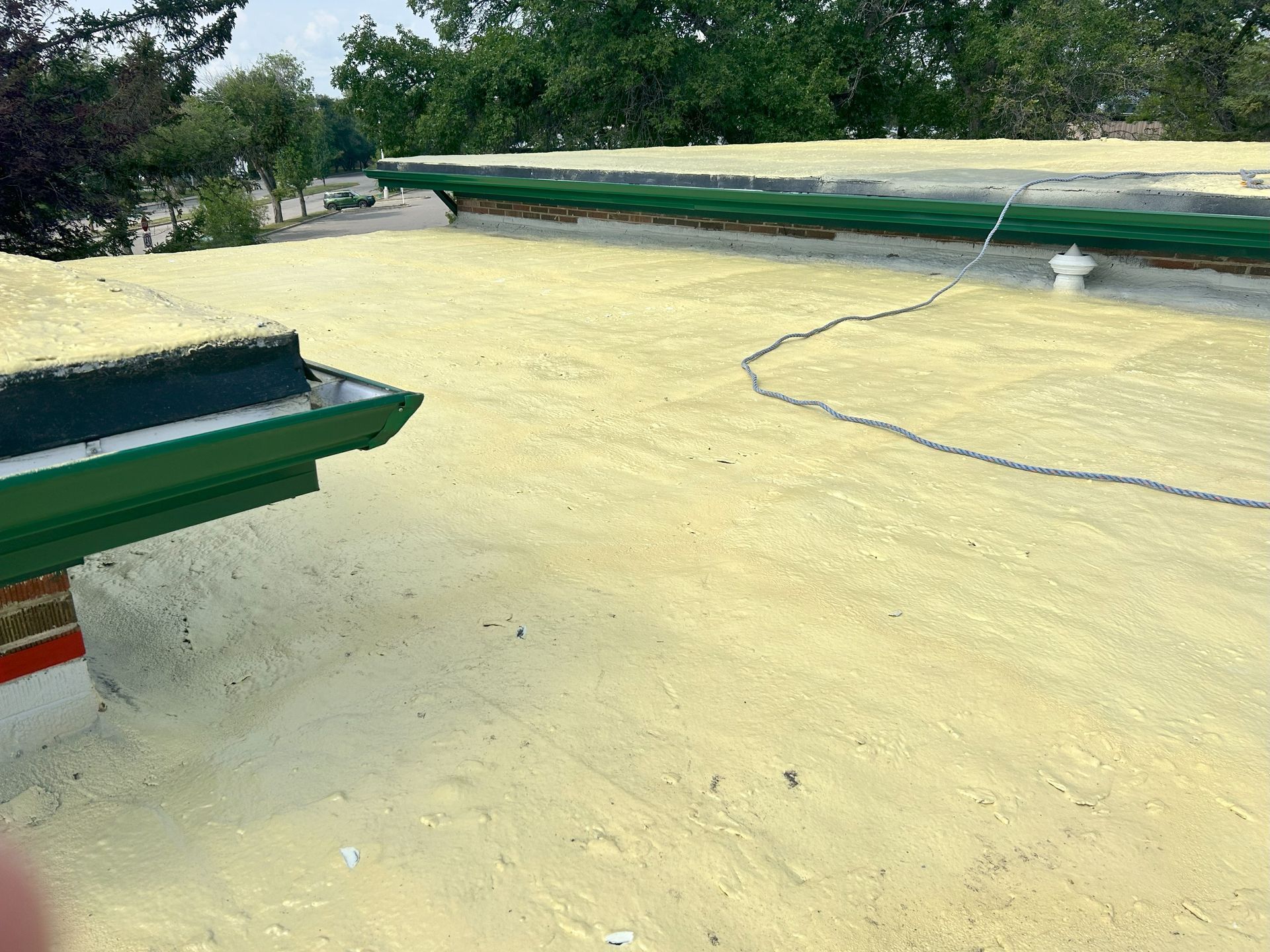 A flat roof with a yellow coating. Green trim and a drainpipe are visible. Trees are in the background.
