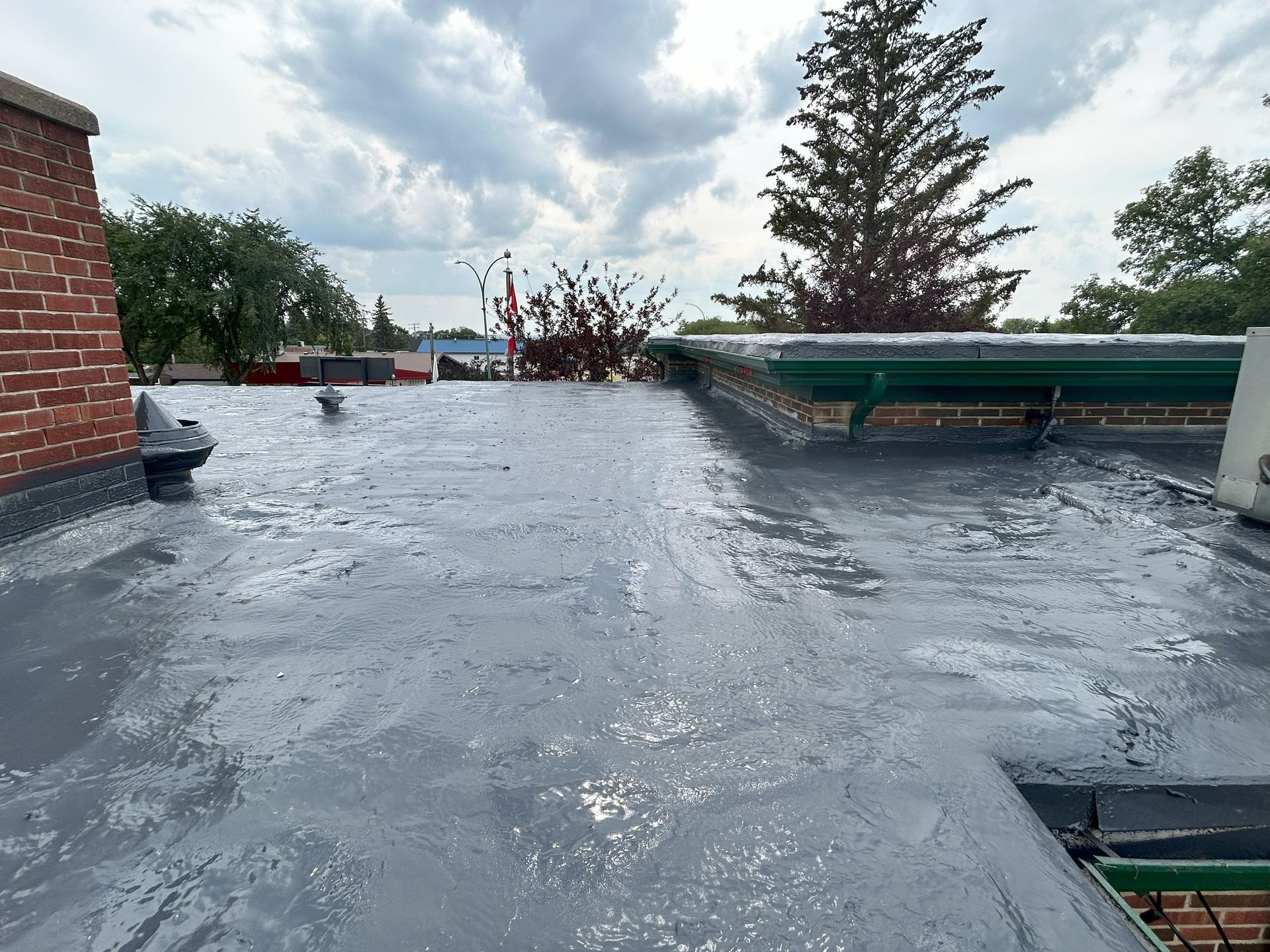 Flat, gray roof with wet coating, against a cloudy sky, with brick buildings and trees in the background.