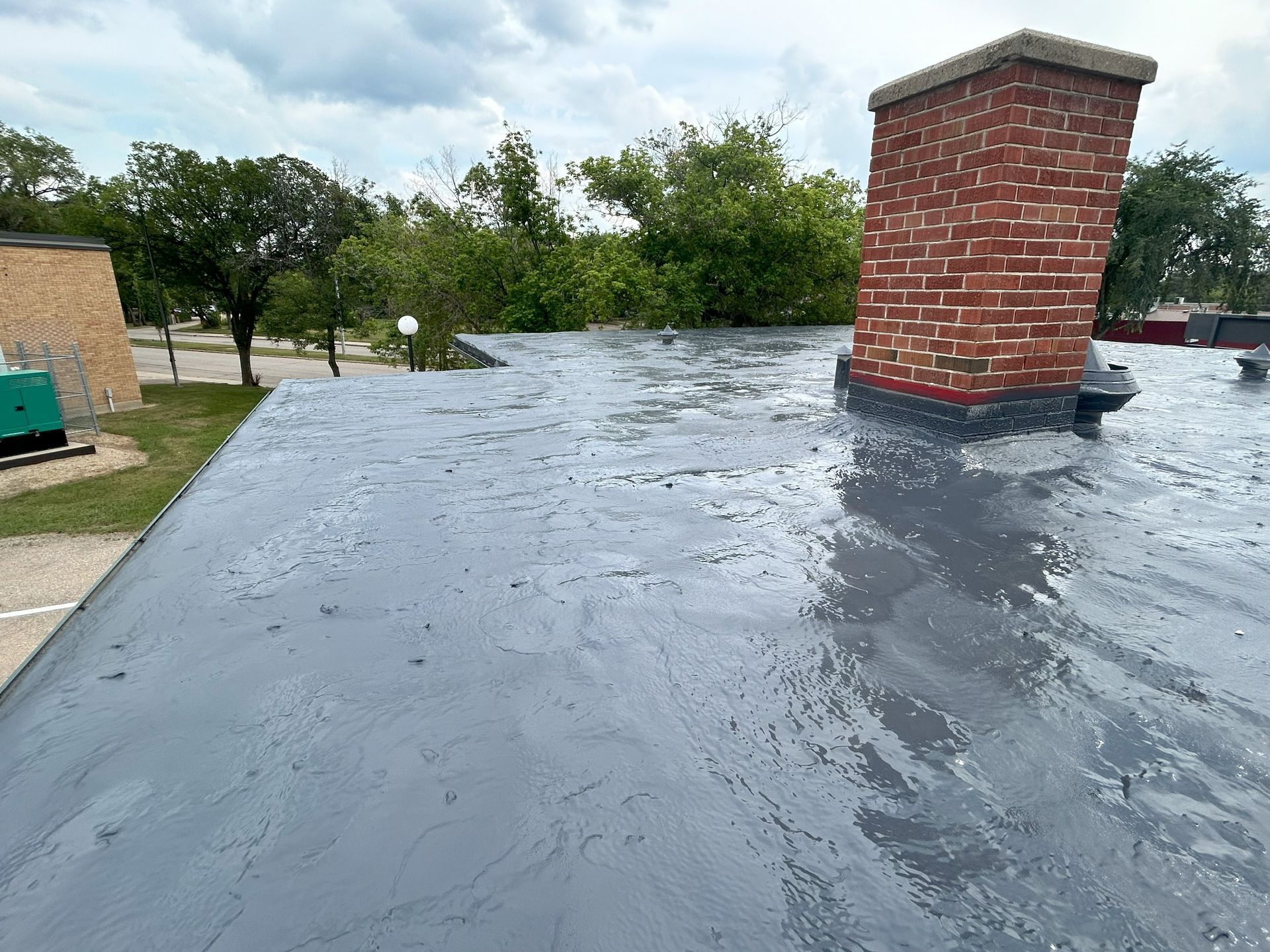 Gray flat roof with a red brick chimney, trees, and a building in the background.