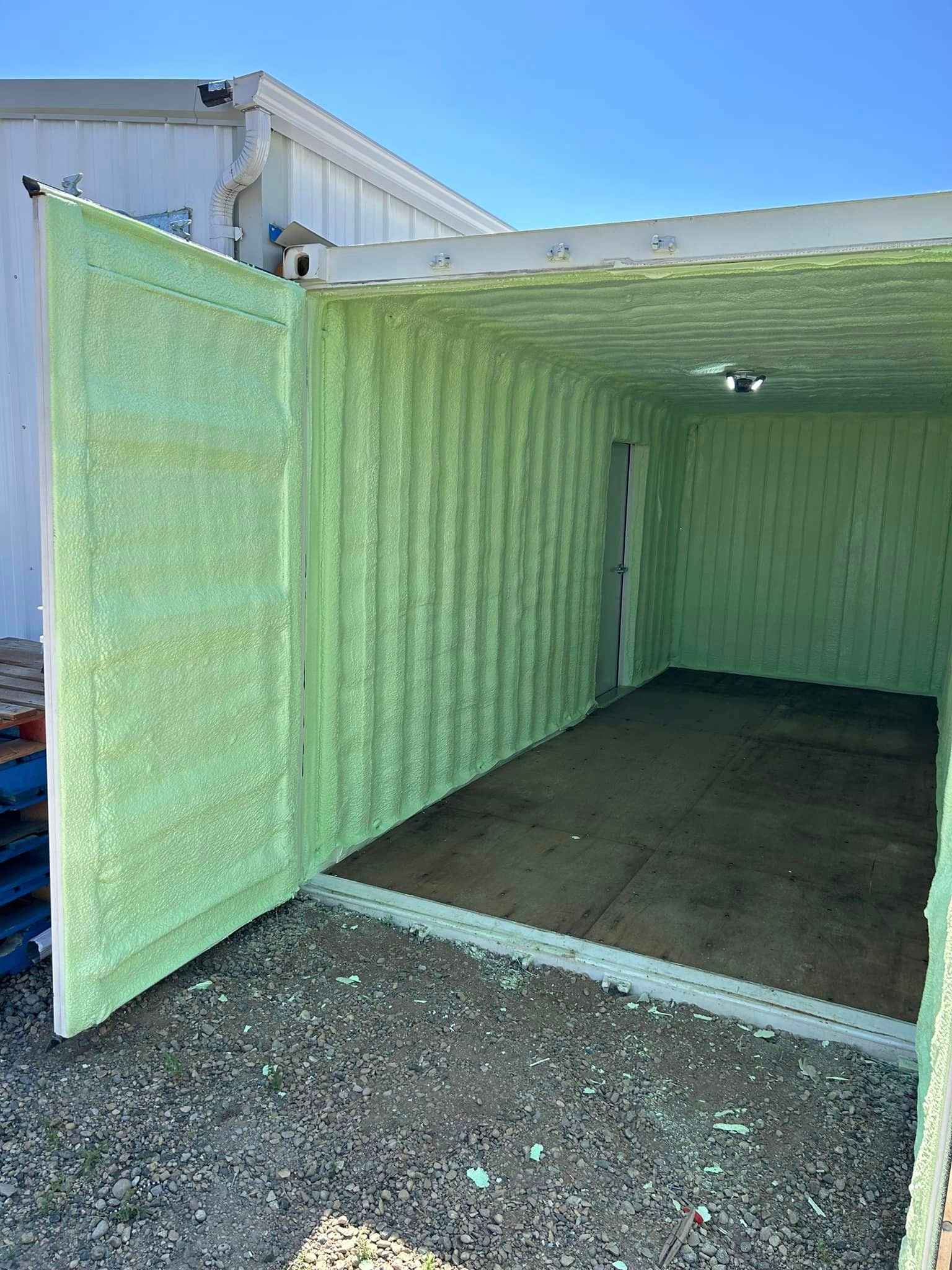 Interior of a container sprayed with green insulation; open door, wooden floor, gravel ground.